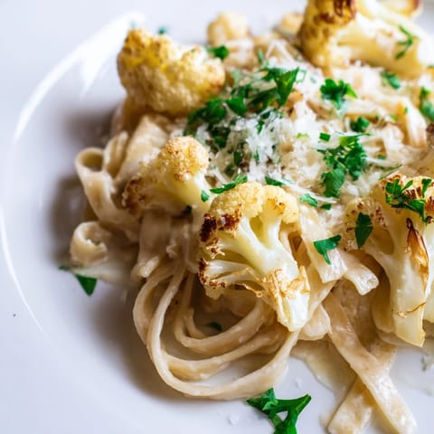 A steaming bowl of Roasted Cauliflower Alfredo, featuring golden-brown florets and creamy sauce clinging to fettuccine, topped with fresh parsley and extra Parmesan.  