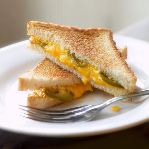 Golden-brown grilled cheese sandwiches filled with melted cheddar and tangy pickled jalapeño slices, served on a rustic cutting board.